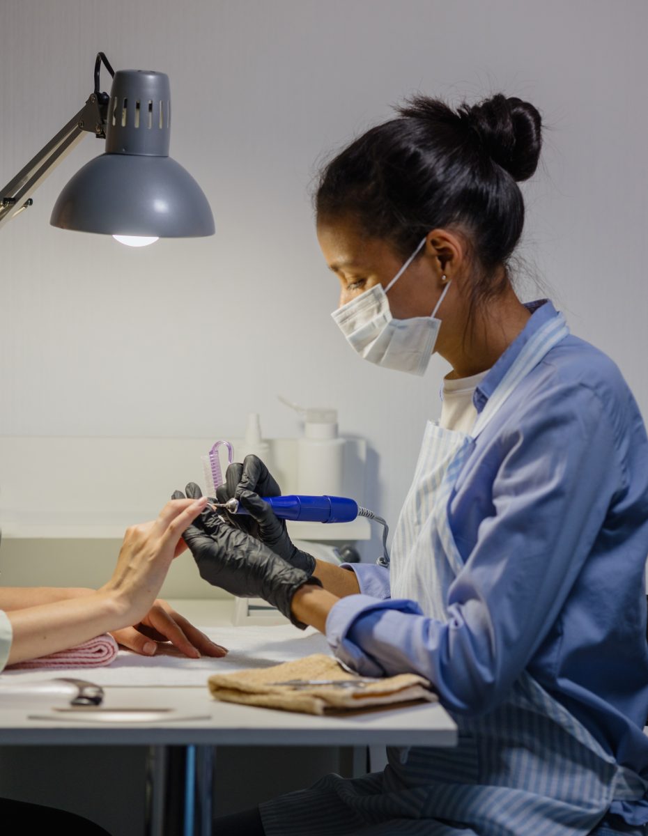 Corded vs. Cordless Nail Drills. An African American woman in a blue shirt and apron doing the client's nails using a nail drill and nail e-file.