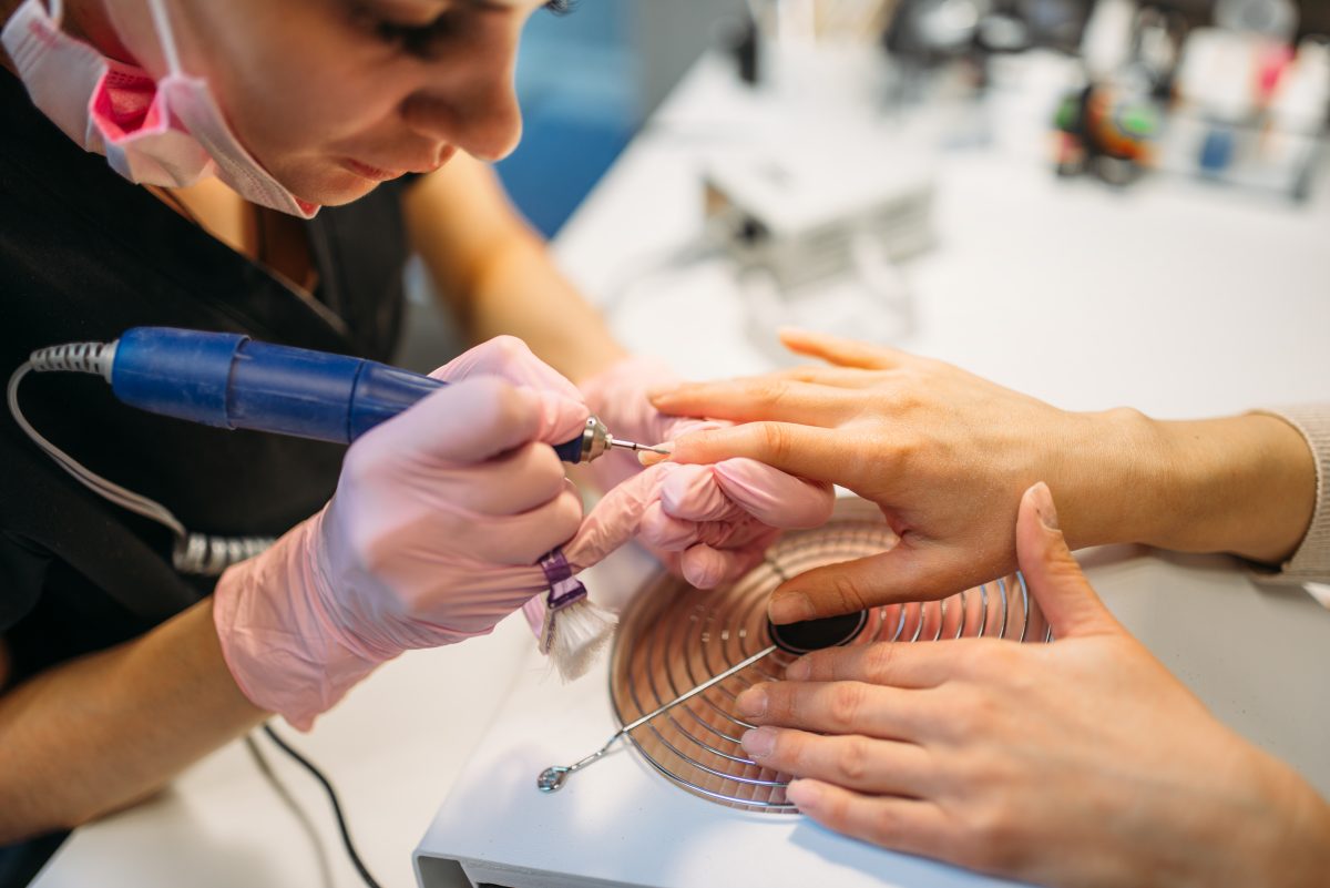 Beautician in gloves with polishing machine treats nails of female client, manicure in beauty salon. Manicurist doing hands care cosmetic procedure