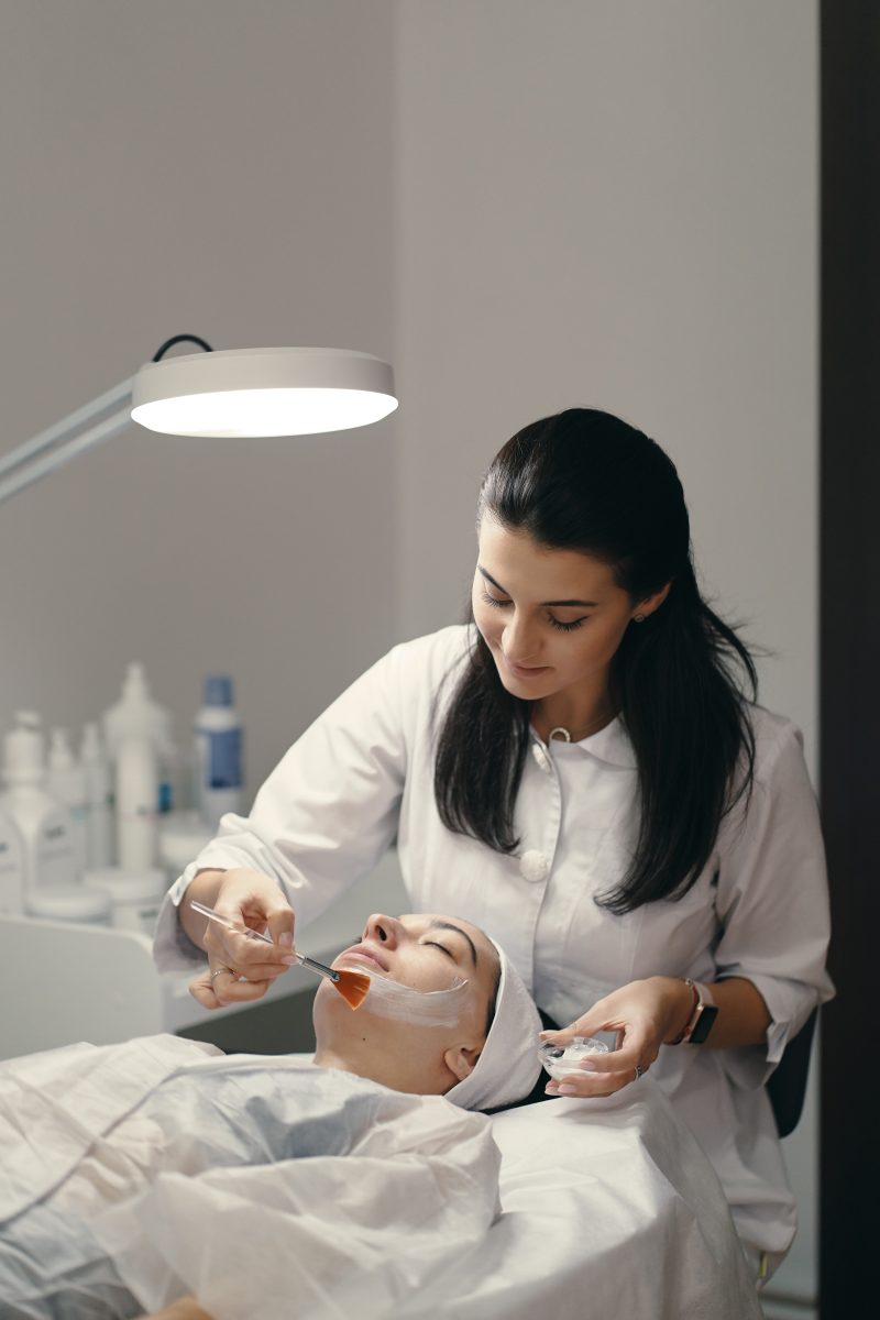 People, beauty, spa, cosmetology and skin care concept - Cosmetologist applying cream on a woman beautiful face with a cosmetologist brush. Lamp light focused on face.