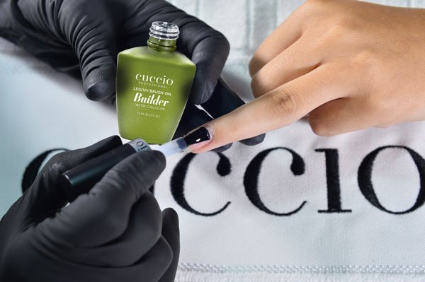 A faceless nail technician wearing black gloves applying Cuccio Brush-On Builder Gel (gel calcium builder) to a client's nails.