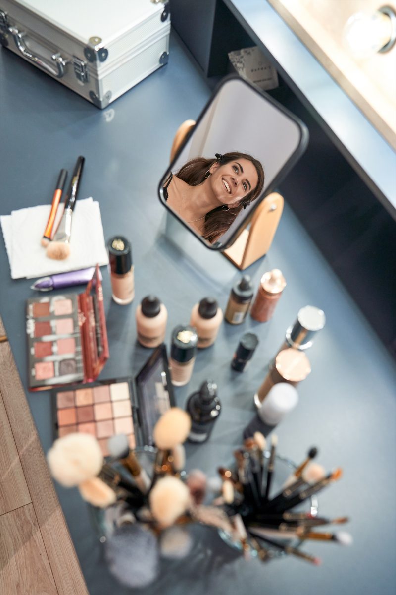 From the above of content, a female client reflecting in the mirror while sitting at a table with a set of makeup supplies in a beauty salon. 