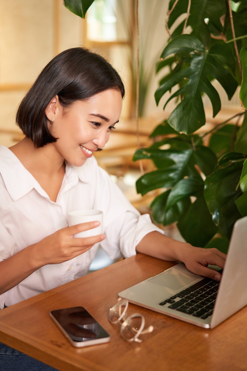 Student sitting in cafe with cup of coffee. Young asian woman attending online beauty course, sitting with computer and smartphone. Beauty online courses.