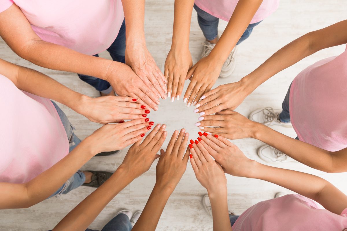 Unrecognizable Diverse Women In Pink T-Shirts Holding Hands In Circle Standing Indoor. Breast Cancer Support. Top View. Different nail shapes and lengths.