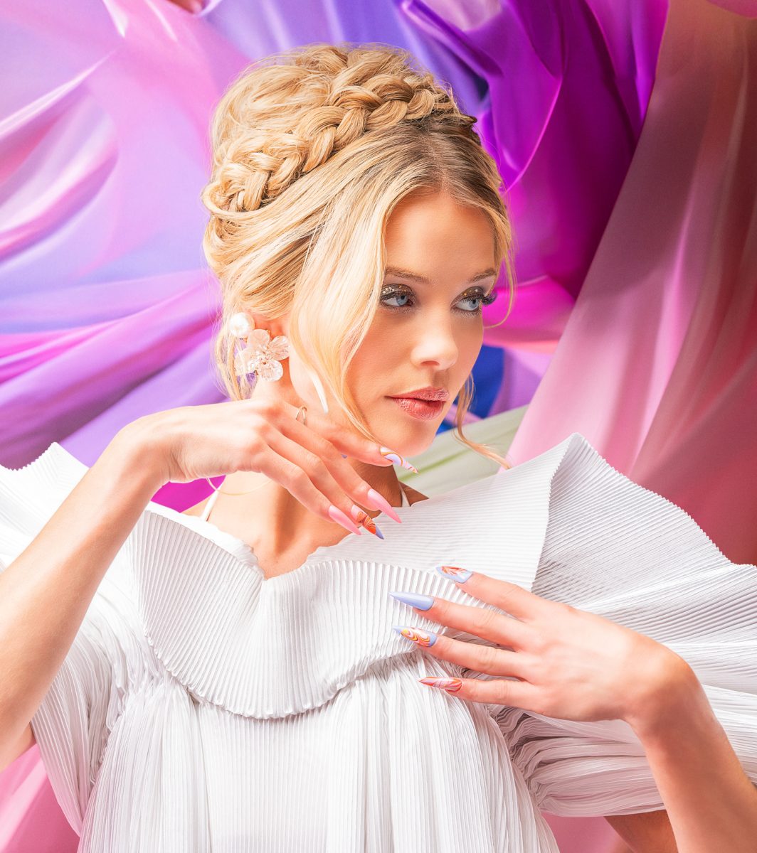A woman with blonde hair styled in an intricate updo is wearing a white textured garment with a large, structured collar. The background features draped fabric in shades of pink and purple. The nails are expertly painted in a pastel color scheme, featuring a mix of soft, serene hues from the Gelish Spring 2025 collection. This image highlights the elegance and sophistication of the season's nail fashion and trends, with the nails serving as a stunning focal point.