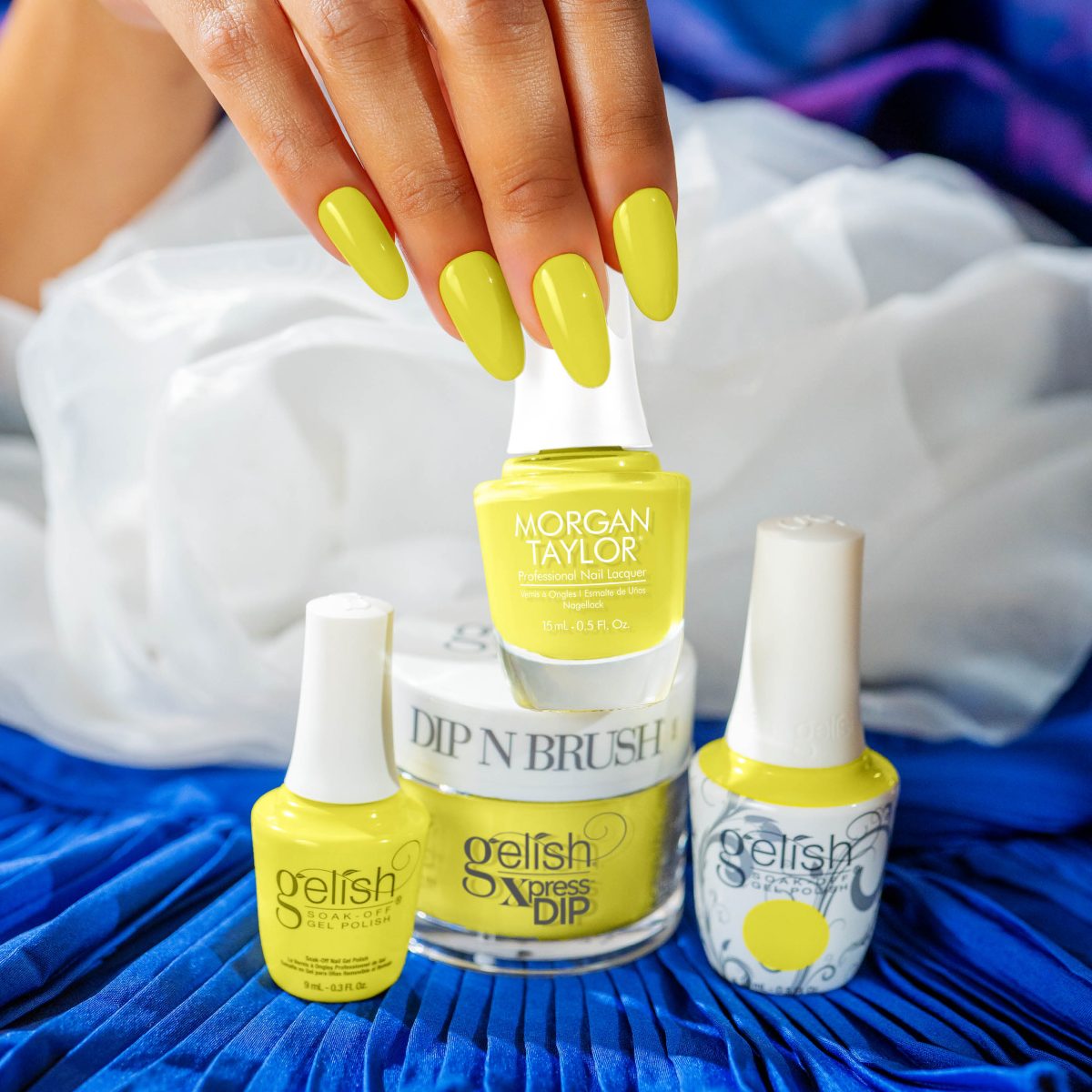 A close-up of a hand showcasing a vibrant yellow nail polish from the Gelish Spring 2025 collection. The nails are beautifully painted in a bright, cheerful yellow shade. In the background, there are three bottles of Gelish nail products, including Gelish Soak-Off Gel Polish, Gelish Dip, and Gelish Dip 'N Brush, all in the same vibrant yellow color. The setting features a white fabric and a blue textured surface, highlighting the colorful and playful nature of the collection.