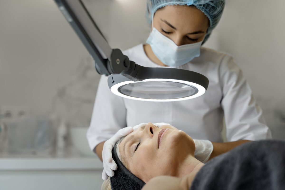 Close up of young masseuse in protective mask massaging mature female patient face. Woman closed eyes with pleasure while laying at the special couch