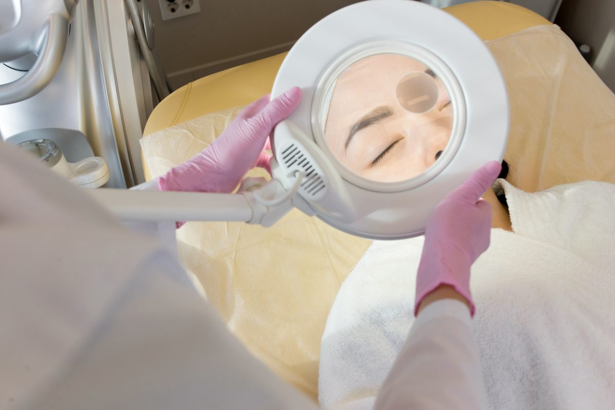 Magnifying Lamps. Close up of unrecognizable cosmetologist inspecting skin of young woman before beauty treatments
