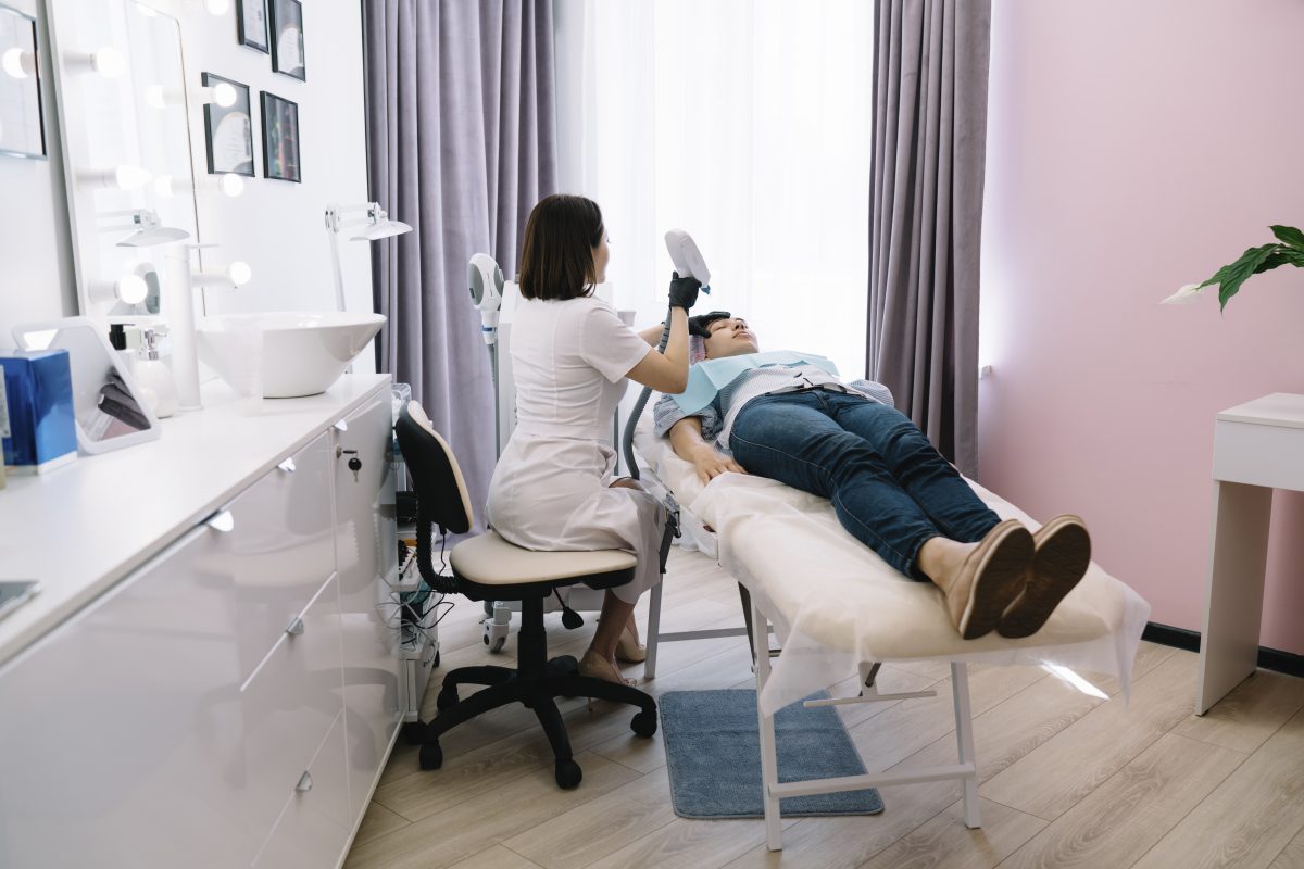 Professional cosmetician in uniform using YAG laser for photo epilation on female clients face lying on couch in modern beauty clinic. Facial Bed + Stool