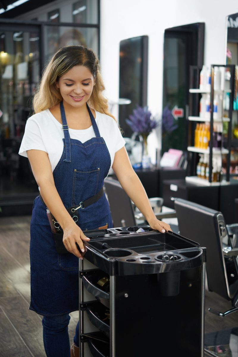 Smiling hairdresser in denim apron pushing storage cart with tools