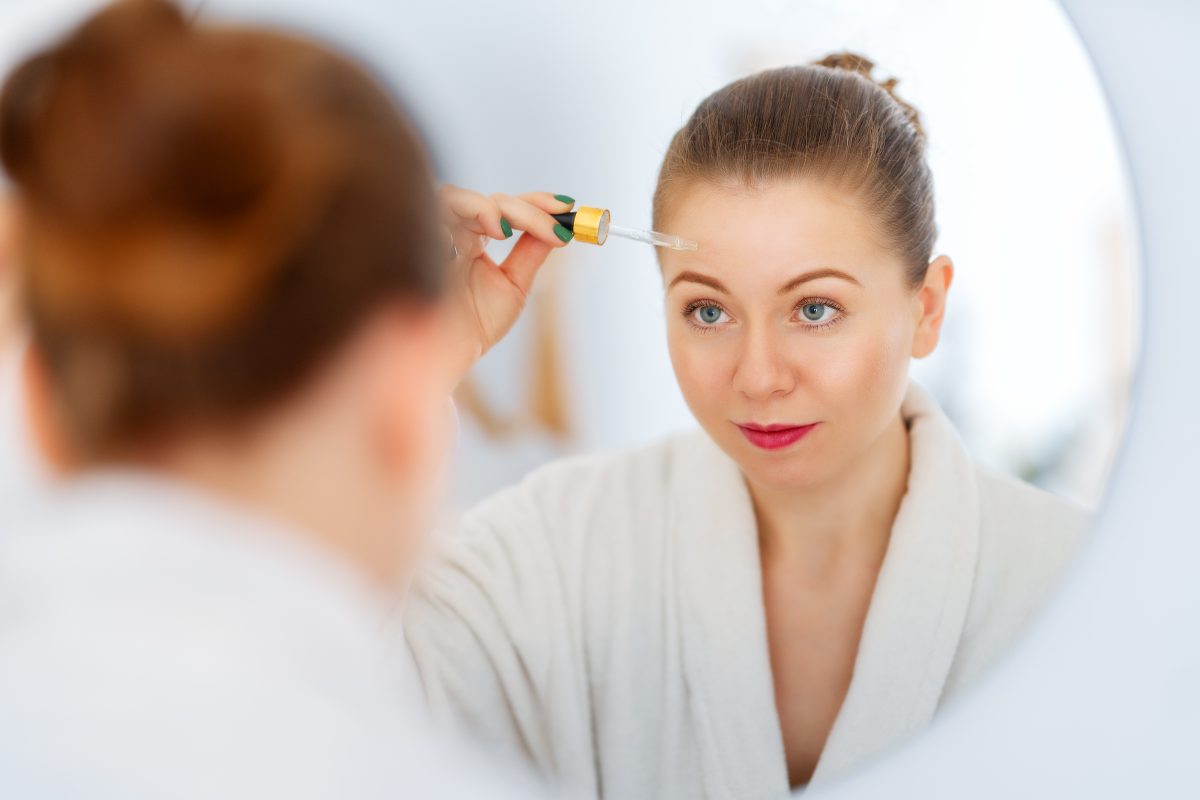 Beautiful young woman with cream for her face looking in the mirror in bathroom at home. Facial treatment. Cosmetology, beauty and spa.