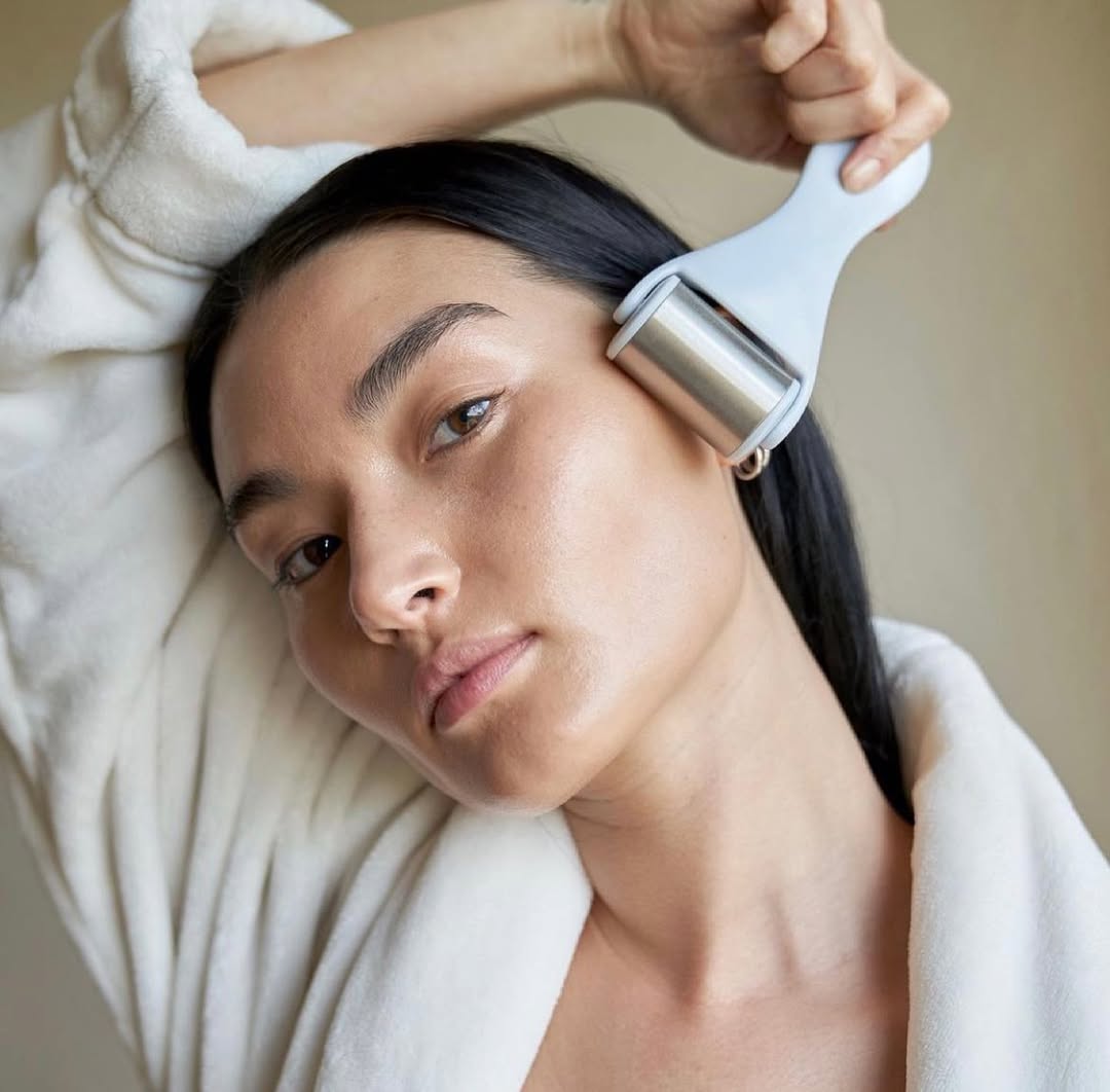 A person in a white robe using a Zoe Ayla ice roller with a metallic roller head and white handle, applying it to the temple area of their face. The background is neutral and softly blurred.