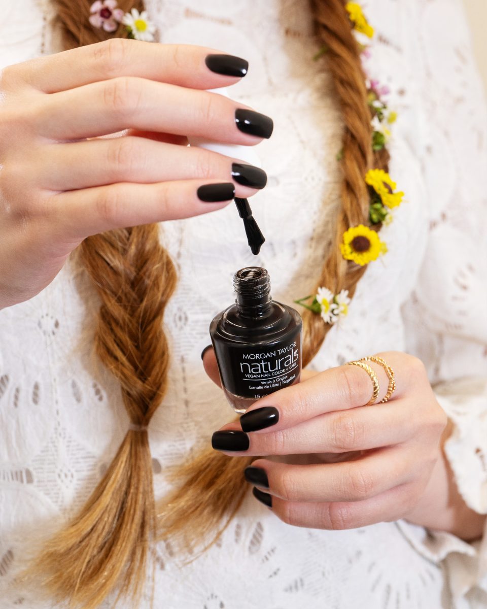 A close-up of a person applying black nail polish from a bottle of Morgan Taylor Naturals. The woman has braided hair adorned with small flowers and is wearing a white lace top. Well-manicured nails using Morgan Taylor Naturals products.