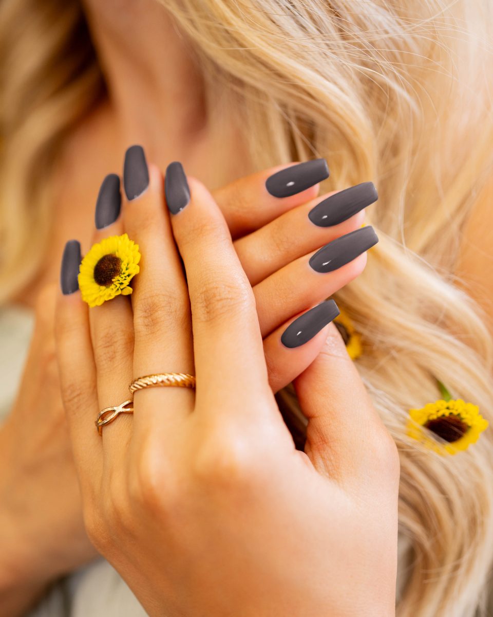 A close-up of hands with long, matte gray nails holding a small sunflower. The hands are adorned with gold rings, and the background features blonde hair with small sunflowers woven into it. Morgan Taylor Naturals nail products.