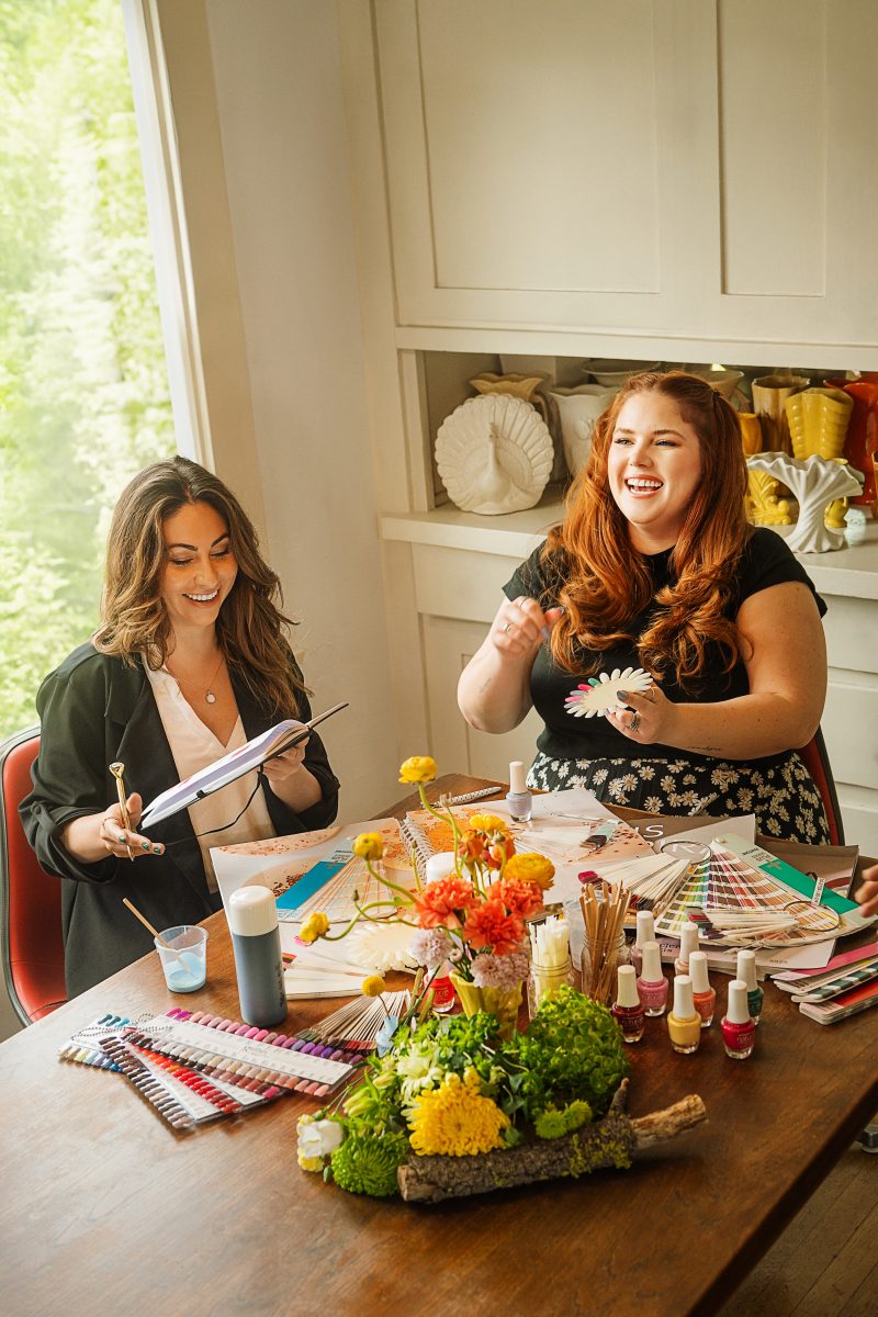 Two women joyfully collaborating on nail color selections at a beautifully decorated table, surrounded by Morgan Taylor Naturals nail polishes, color swatches, and floral arrangements.