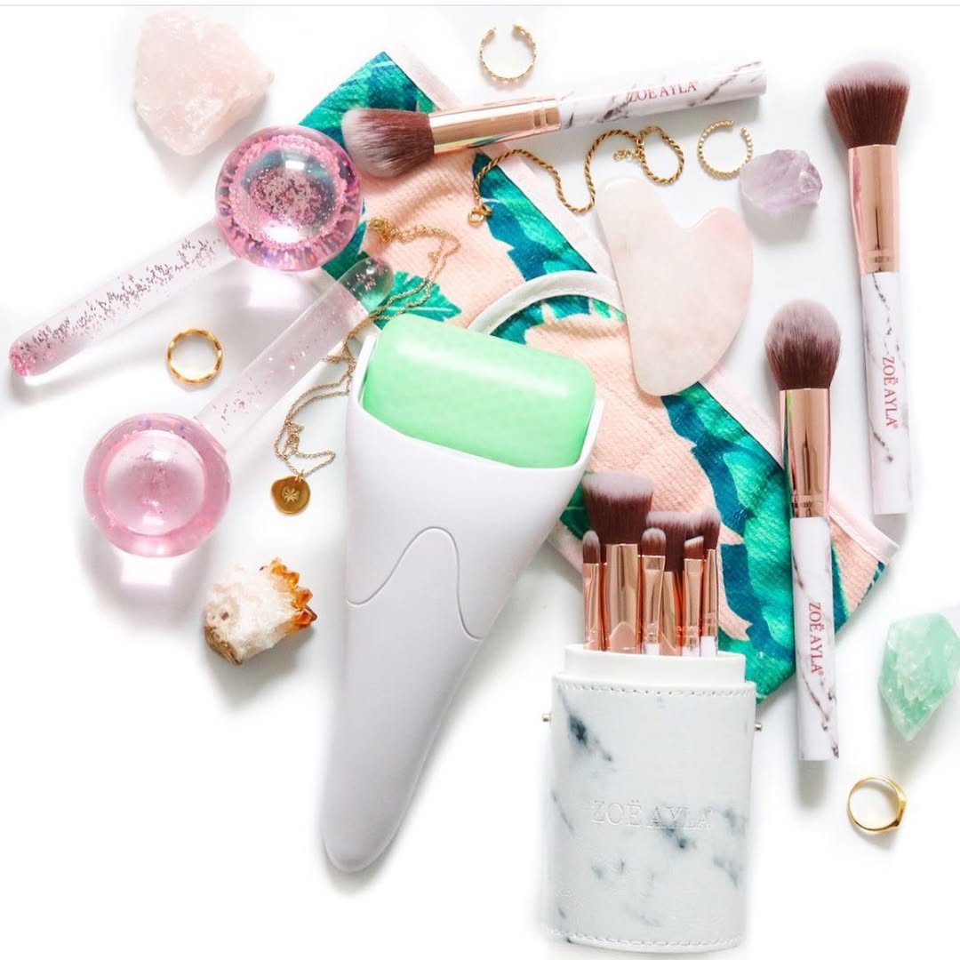 A stylish arrangement of Zoe Ayla beauty tools and accessories displayed on a sleek white surface. The collection includes makeup brushes with elegant white and rose gold handles, a white marble-patterned brush holder, a pink facial roller, a matching Gua Sha tool, an ice roller, and various decorative items like crystals and jewelry. The layout emphasizes the brand's luxurious yet functional aesthetic, blending modern design with high-quality craftsmanship.