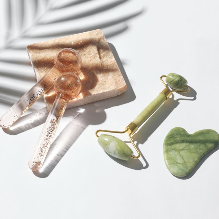 A beautifully arranged set of Zoë Ayla skincare tools displayed on a sleek white surface. The arrangement includes two elegant glass globes filled with liquid and gold flakes, a green jade facial roller with gold accents, and a matching green jade Gua Sha tool. The tools are artfully positioned, with the globes placed on a beige stone block and the roller and Gua Sha tool resting directly on the surface. A shadow of a palm leaf softly falls in the background, adding a natural and luxurious touch to the composition.