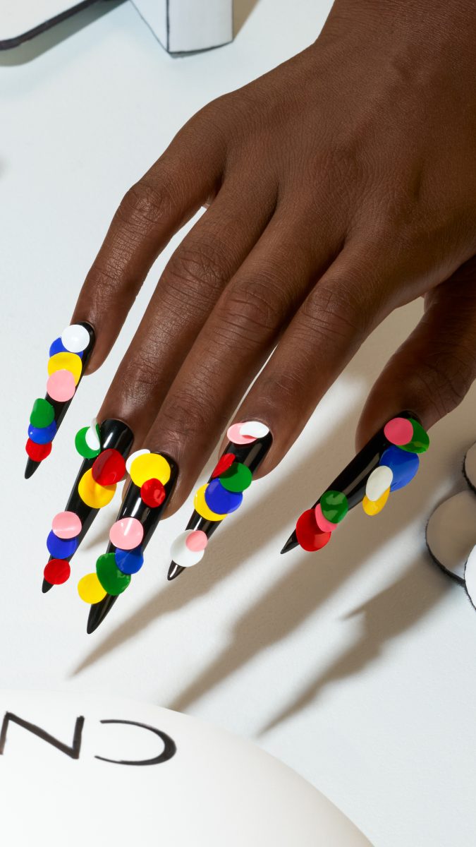 A hand showcasing long, pointed black nails with vibrant and colorful circular patterns. The design features an array of colors like red, yellow, green, blue, pink, and white, creating a playful and eye-catching look from the CND Summer 2025 collection: SHELLAC™ VIVID.