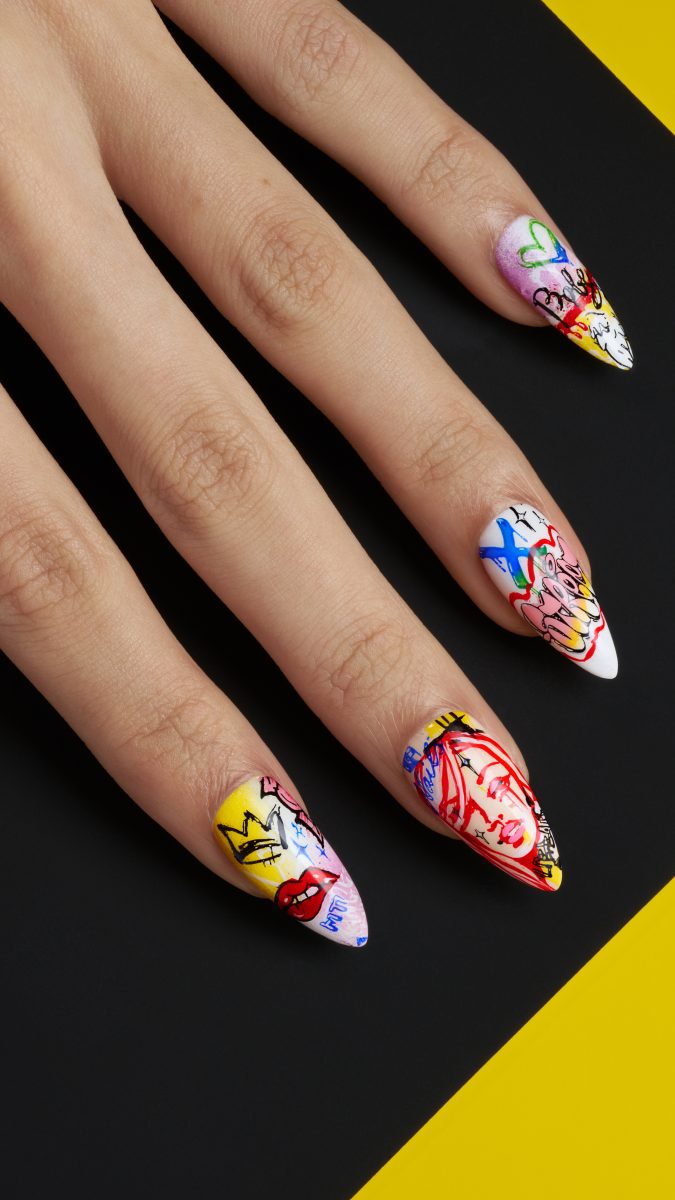 A vibrant display of nail designs from the CND Summer 2025 collection: SHELLAC™ VIVID. The nails are long and pointed, featuring unique and colorful patterns, including abstract shapes, graffiti-style art, and doodles with shades of yellow, red, blue, white, and more. Elements such as lips, crowns, and playful icons create an energetic and artistic vibe. The black background makes the vivid colors and intricate designs stand out.