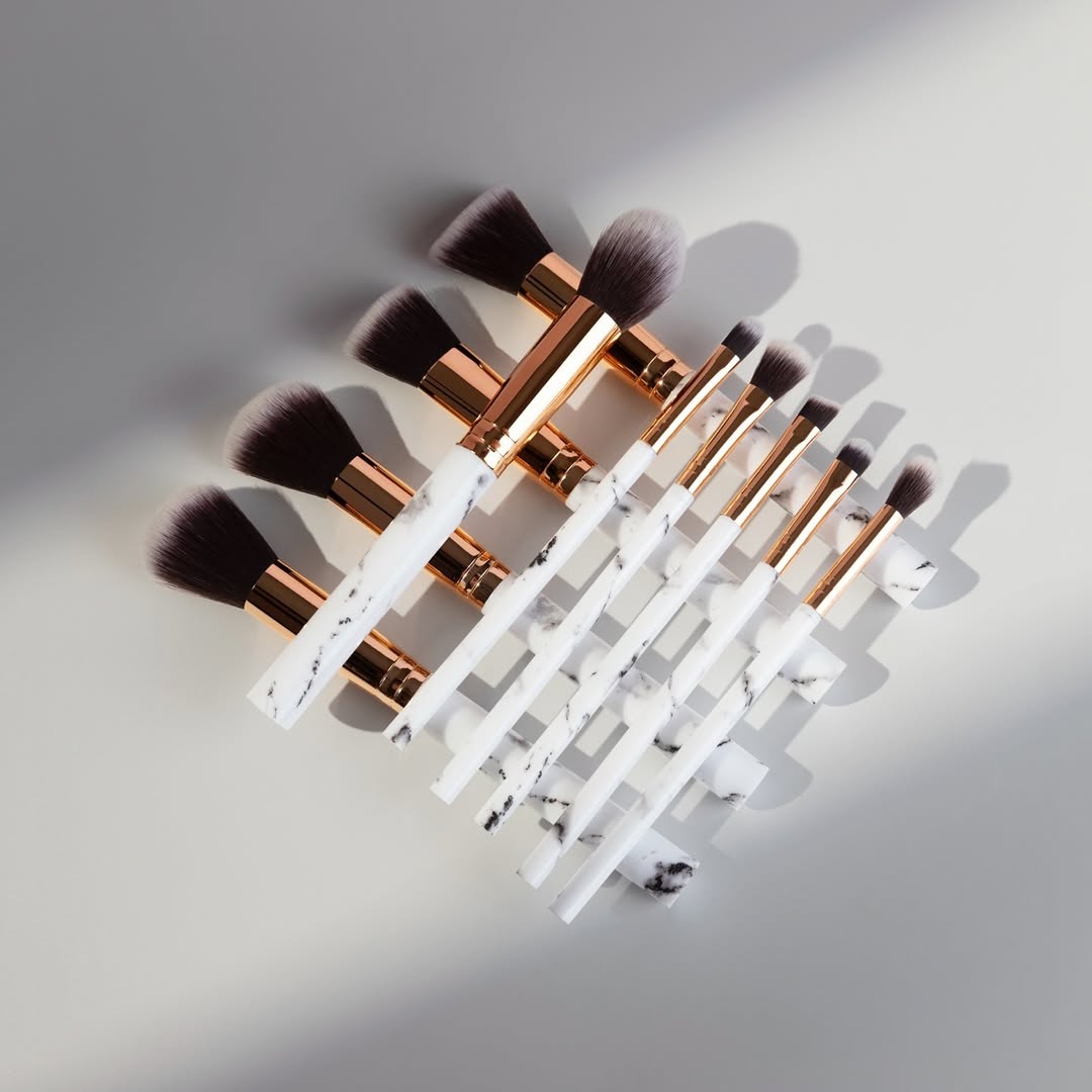 A set of Zoe Ayla makeup brushes laid out neatly on a light-colored surface. The brushes feature white handles with a sophisticated black marble pattern and rose gold ferrules. The top row consists of five large, fluffy brushes, while the bottom row includes five smaller brushes with fine bristles. The arrangement forms a visually appealing display with soft shadows accentuating their elegant design.