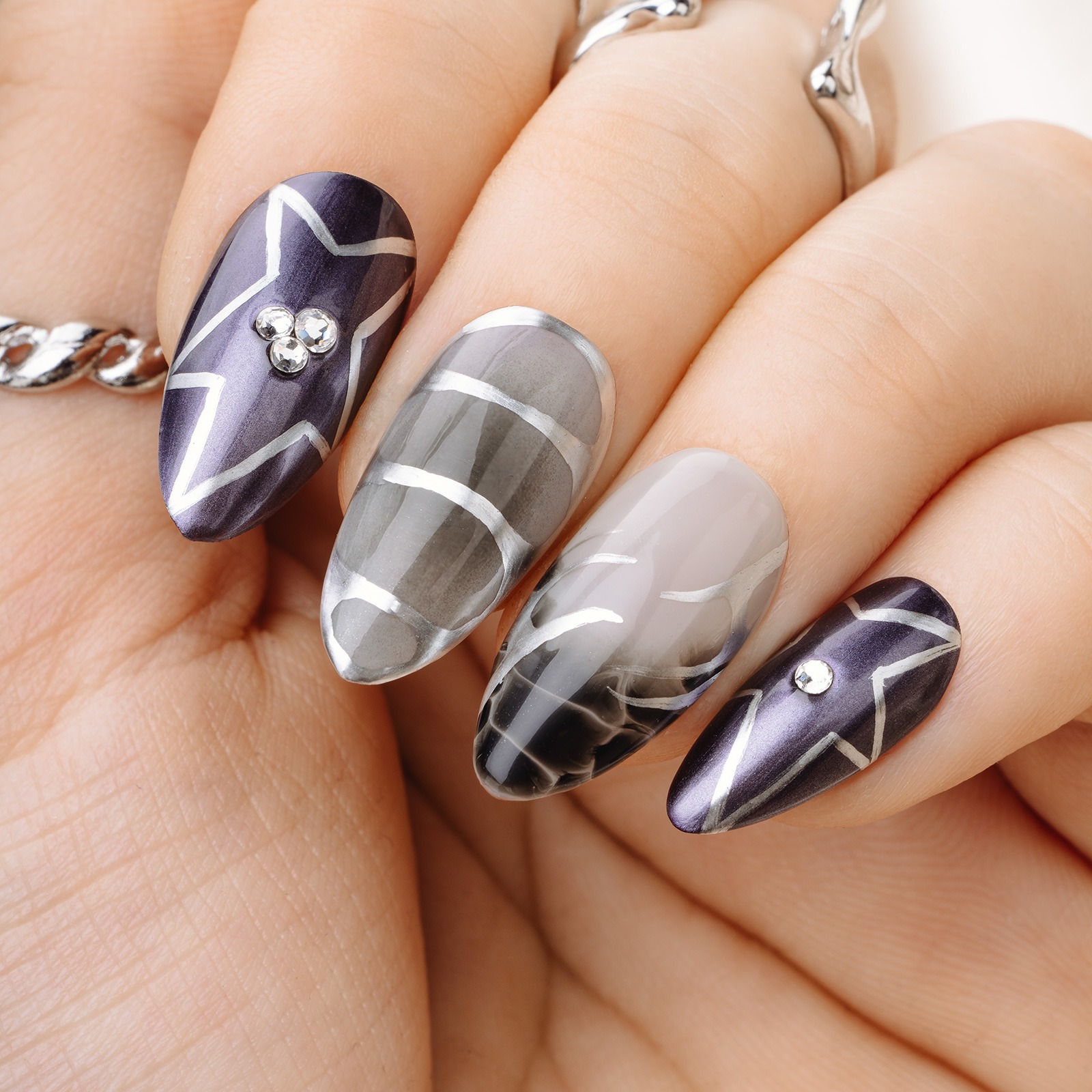 A close-up of a hand adorned with long, almond-shaped nails showcasing intricate nail art created with Apres Silk Cover. The design features dark purple tones accented with silver stars and rhinestones on the thumb and pinky. The index and middle nails display a gradient effect in gray and white with abstract silver stripes. The hand is accessorized with elegant silver rings and a chain bracelet, all set against a neutral background. This highlights the creativity and versatility of the Apres Silk Cover Collection