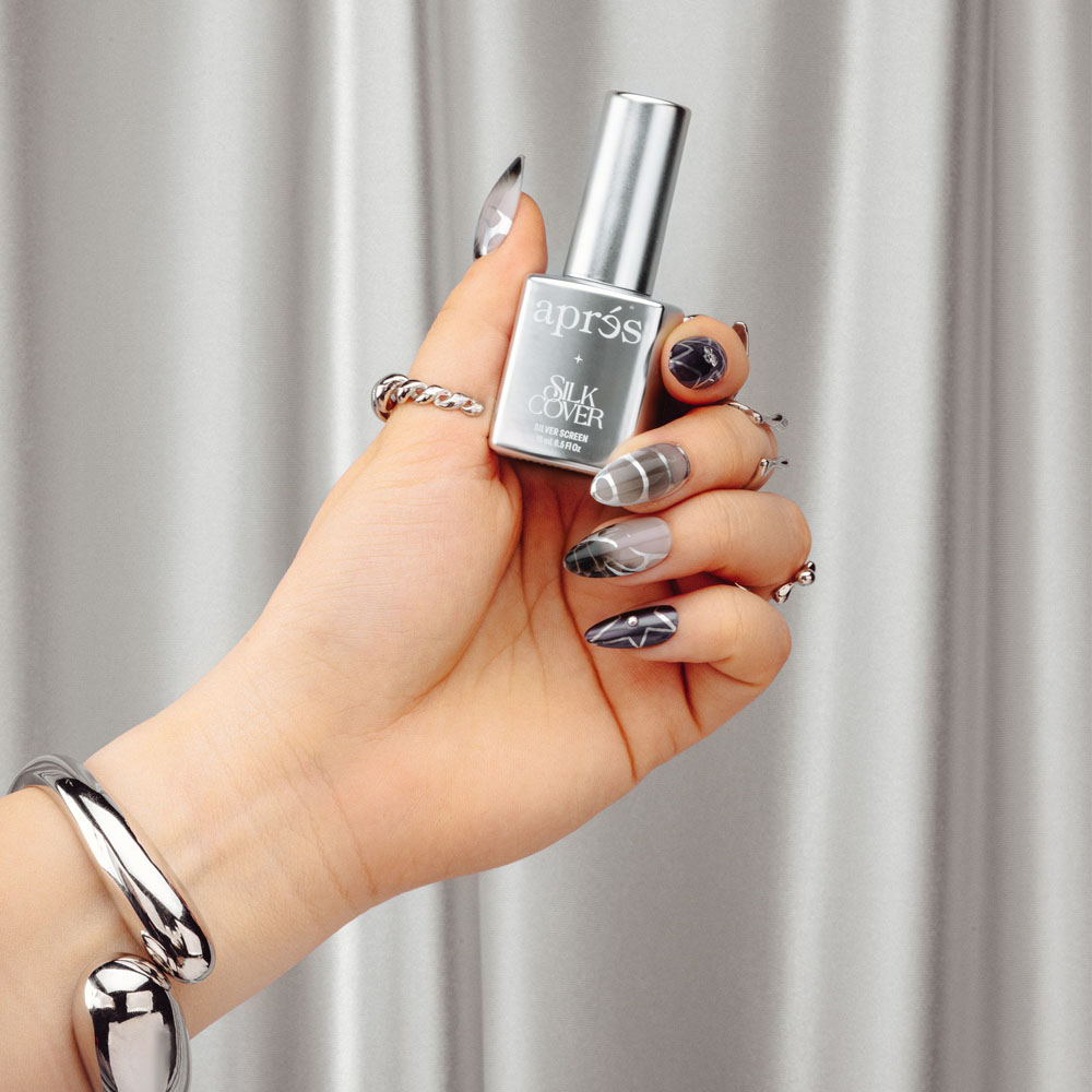 A hand with manicured nails holds a bottle of "après Silk Cover Silver Screen" nail polish. The nails showcase intricate designs, blending geometric and abstract patterns in black, white, and silver. The hand is adorned with silver rings and a bracelet, set against a textured silver metallic backdrop.