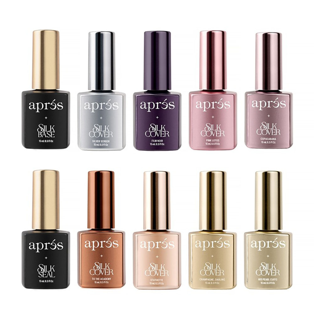 Ten bottles of Apres Silk Cover nail polish arranged neatly in two rows of five on a clean surface. The top row includes shades such as Silk Base, Silver Screen, Film Noir, Pink Lotus, and Copacabana, while the bottom row features Silk Seal, To The Academy, Statuette, Champagne, Darling, and His Pearl Cuffs. Each bottle is labeled with its name and features a consistent, sleek design. The colors range from metallic silver and gold to vibrant pink and sophisticated black, showcasing the luxurious and professional-grade collection.