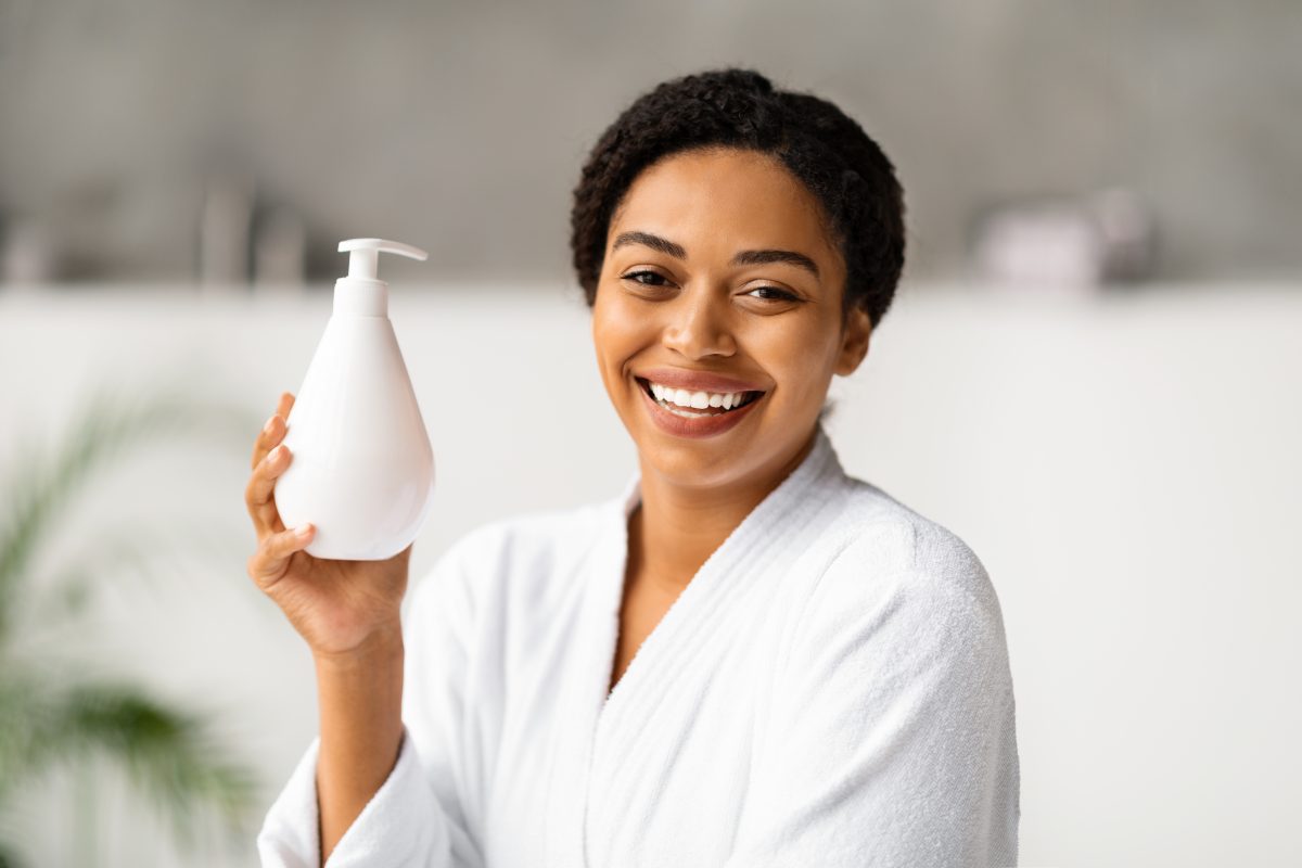Body Butters vs Lotions Understanding the Key Differences. Beautiful Smiling African American Woman In Bathrobe Holding Bottle With Moisturising Body Lotion, Happy Black Lady Using New Skincare Product After Bath, Standing In Modern Bathroom Interior.