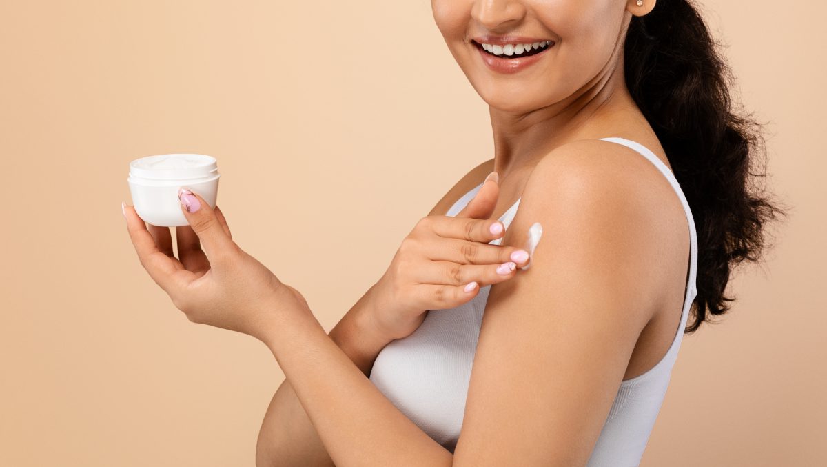 Beautiful Young Woman Applying Moisturizing Cream On Shoulder, Closeup Shot Of Smiling Indian Female Nourishing Skin While Standing On Beige Studio Background, Holding Jar With Skincare Cosmetics. Vegan body creams. Body cream. 