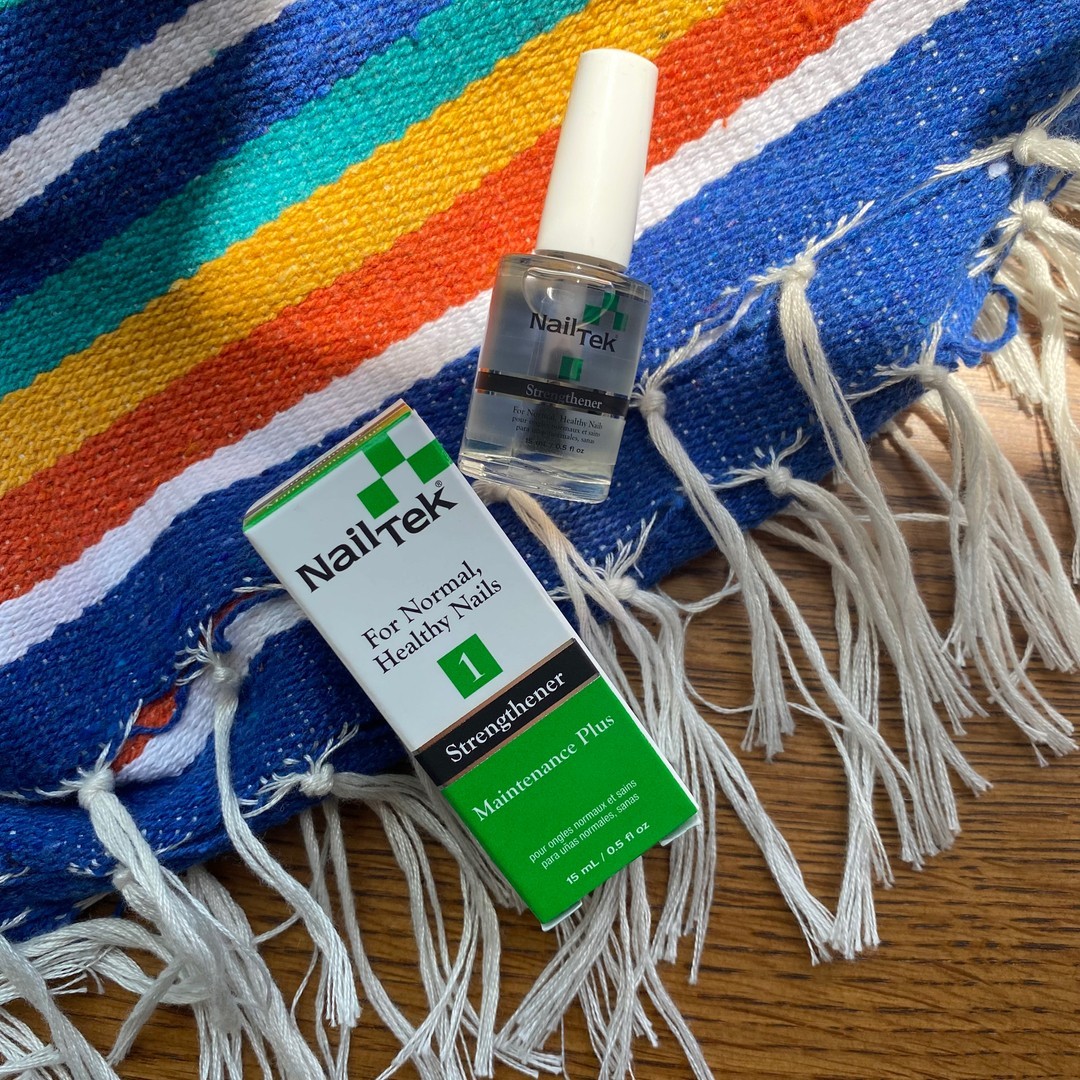 Maintenance Plus 1 Strengthener as a base coat will help condition, protect and maintain normal, healthy nails. A bottle of Nail Tek nail strengthener alongside its packaging, set on a vibrant woven fabric with fringe details. The packaging displays the label "Nail Tek" and specifies the product as "1 Strengthener Maintenance Plus," designed for normal, healthy nails. The clear bottle with a white cap bears the Nail Tek logo, complementing the professional and visually appealing presentation.