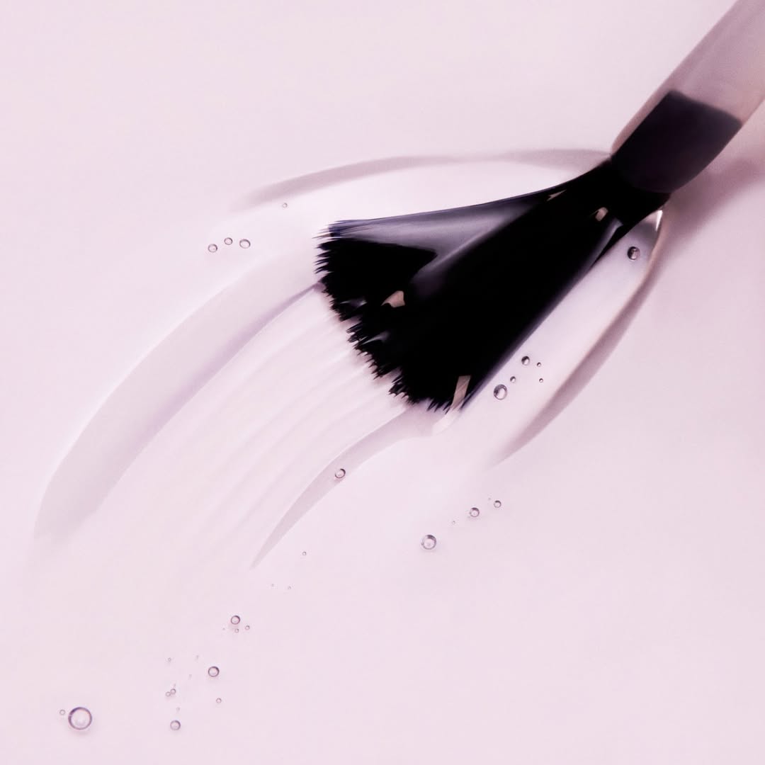 A detailed close-up image of a Nail Tek product brush with sleek black bristles. The brush is shown gliding smoothly on a light pink surface, creating a delicate stroke of liquid product. Surrounding the brush stroke, small droplets reflect the active application process, emphasizing the precision and effectiveness of the tool.