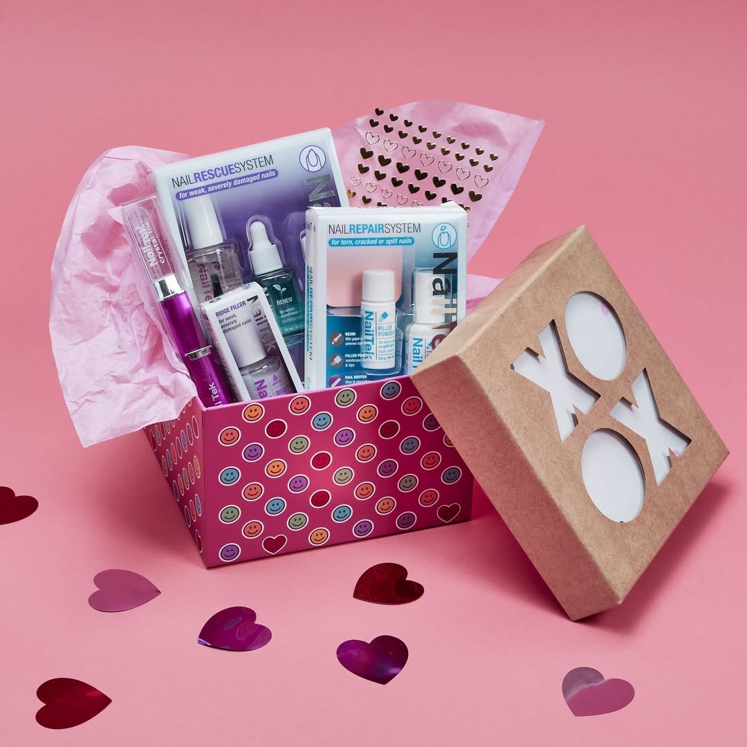 A vibrant presentation of Nail Tek products inside a pink gift box with a colorful circular pattern. The box includes a personalized nail treatment tailored to your nail type, a Nail Repair Kit, a set of playful nail stickers, Ridge Filler, and a Crystal Nail File. The box lid, placed beside it, features "XOXO" cut-out letters. The background is pink with heart-shaped confetti scattered around, creating a cheerful and elegant display.