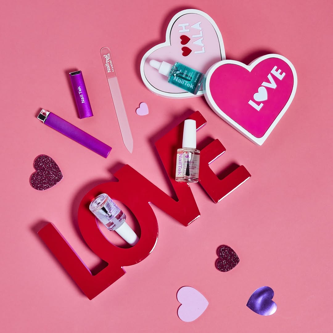 A visually appealing arrangement of Nail Tek products displayed on a pink background adorned with heart-shaped decorations. The centerpiece features a bold red "LOVE" sign, surrounded by bottles of Nail Tek Foundation and Nail Tek Intensive Therapy. Additional items include a nail file, a purple lighter, and a heart-shaped container labeled "LOVE," creating a theme of self-care and affection. The setup exudes warmth and elegance, emphasizing nail care and beauty.