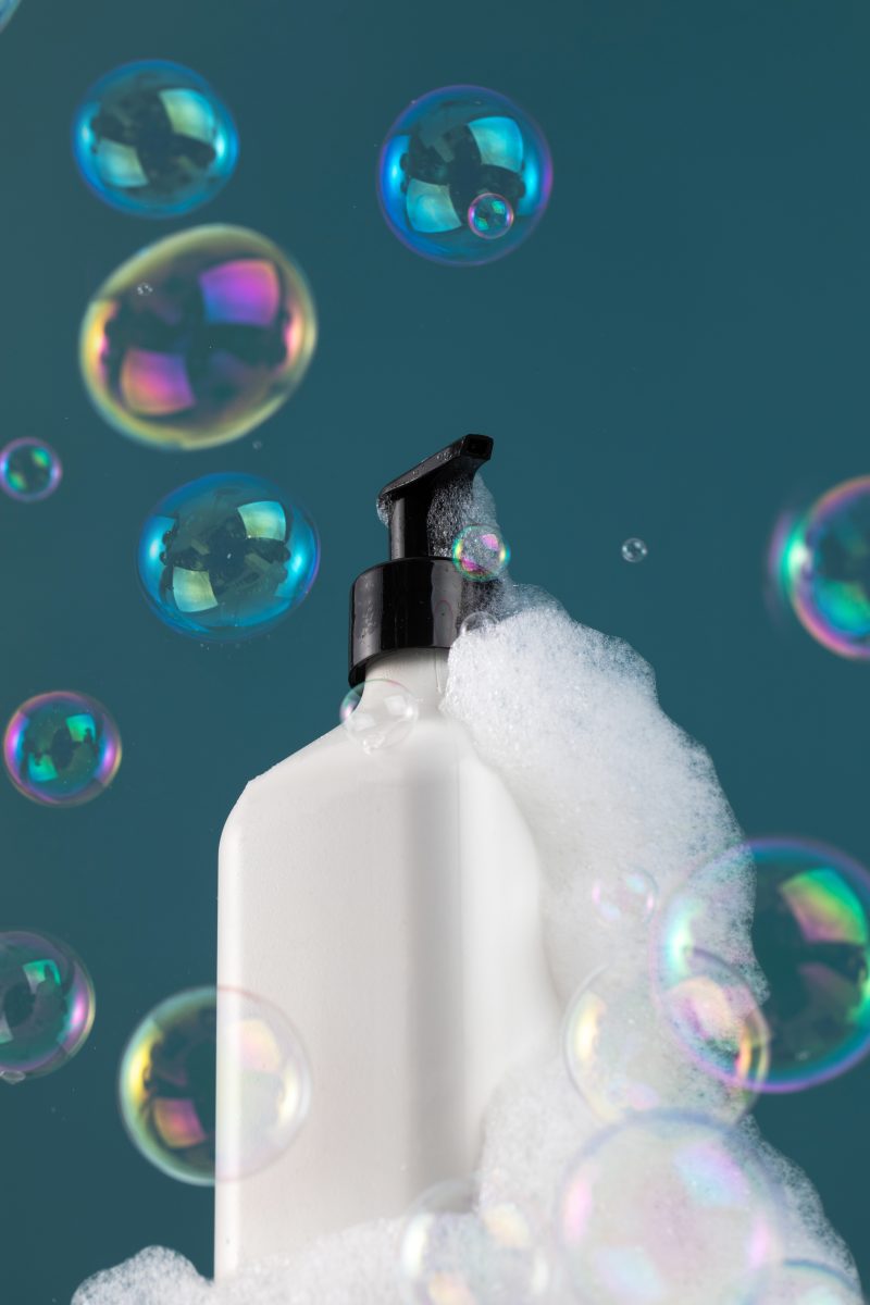 A white dispenser with a cosmetic product in foam on the shelf and with flying soap bubbles. The concept of shower gel, shampoo. What to Expect When Switching to Sulfate-Free Shampoo.