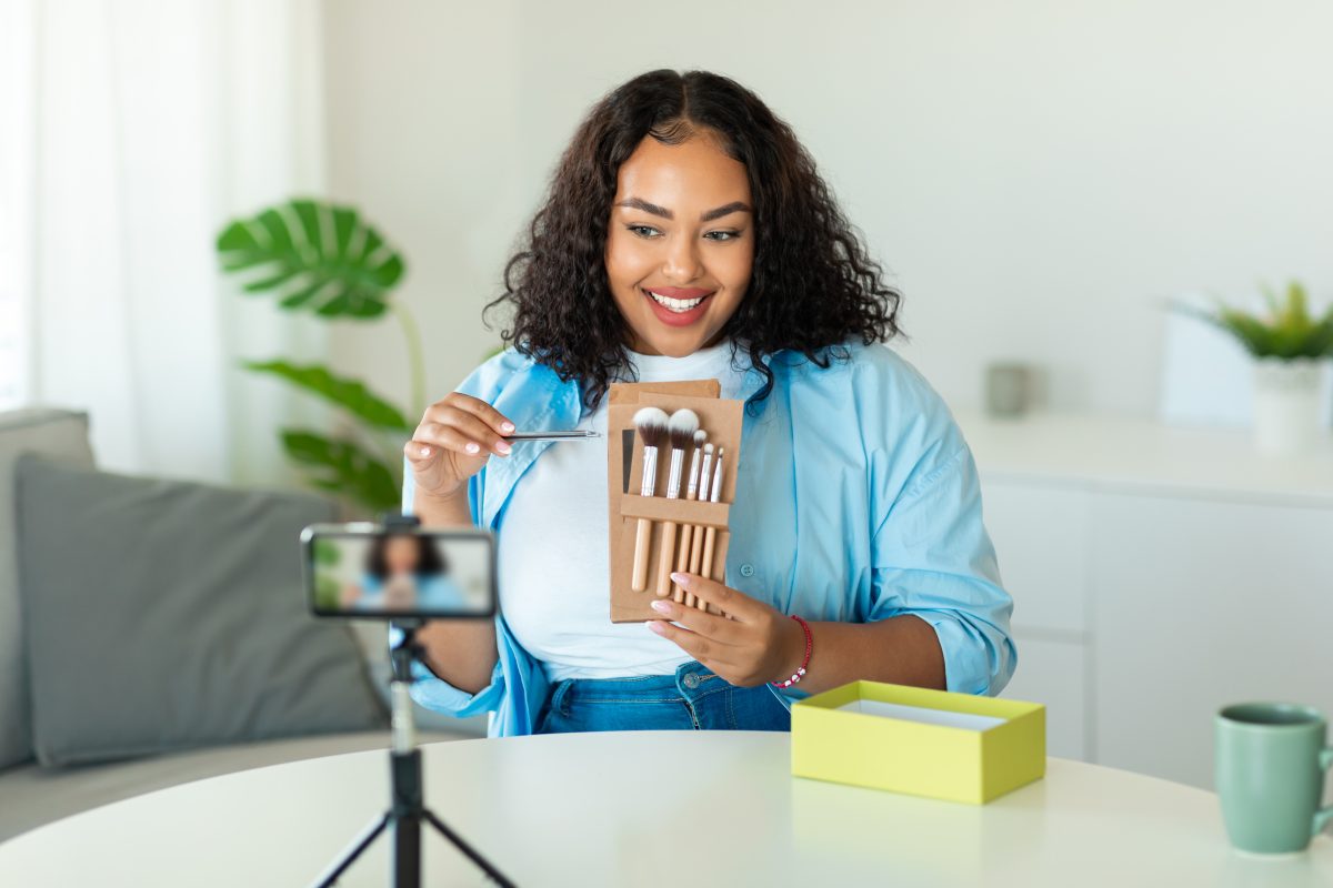 Beauty Blogging. Black Blogger Lady Making Makeup Video Tutorial For Her Blog Filming On Cellphone And Showing Cosmetic Brushes Sitting At Desk At Home. Selective Focus