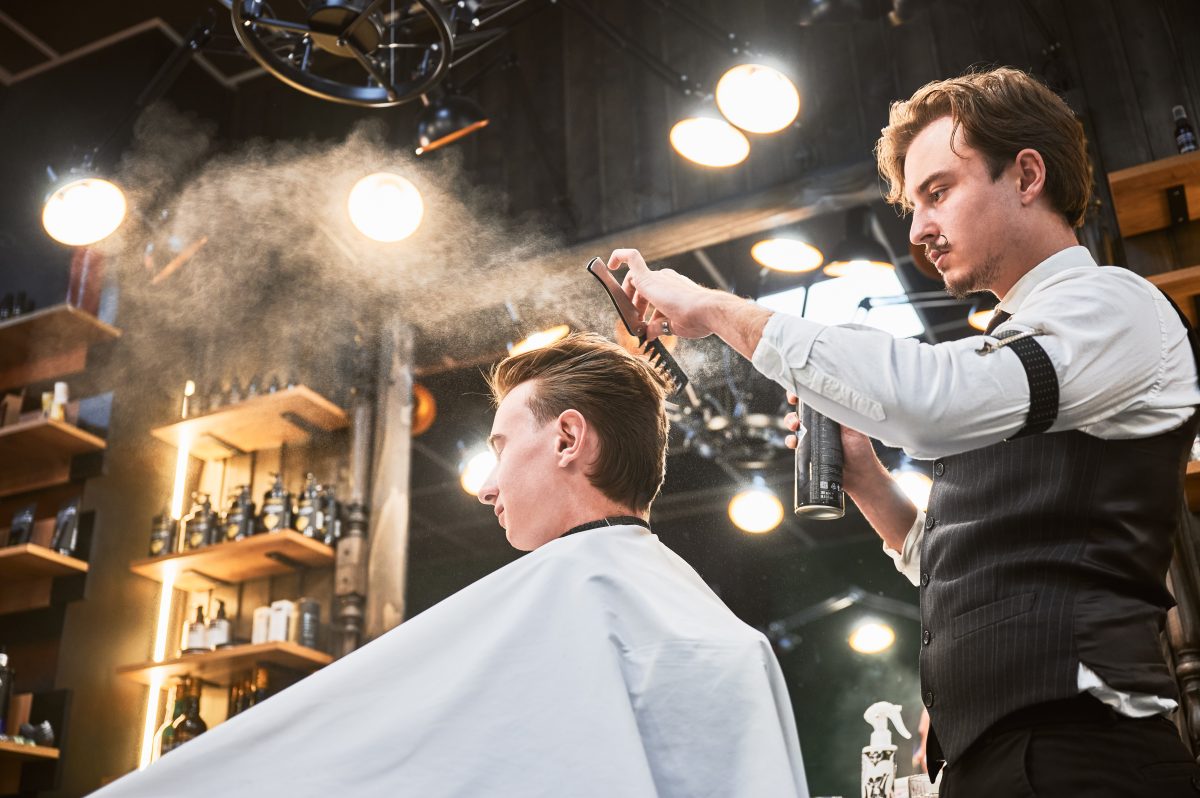 Professional barber using hairspray to finish hairstyle. Stylish hairdresser making final touches. Young customer getting fresh haircut.