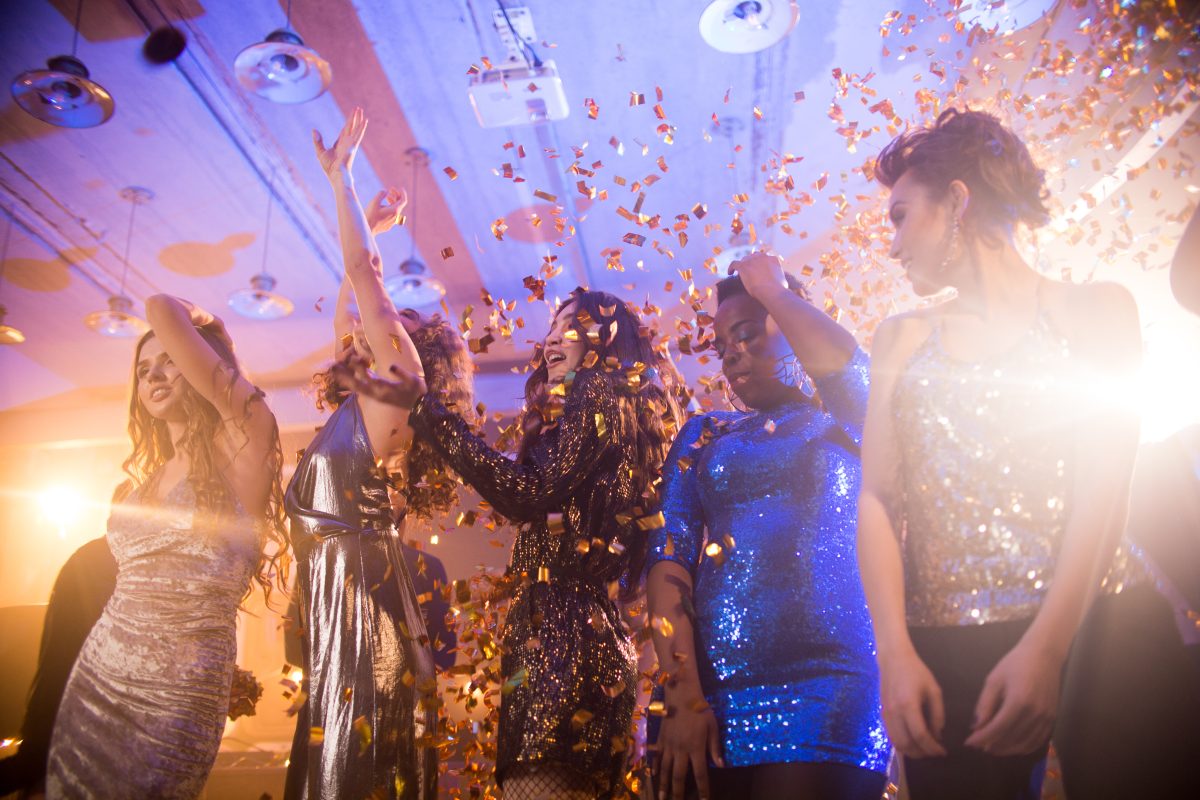 Group of beautiful young women wearing glittering dresses dancing under golden confetti shower enjoying party in nightclub