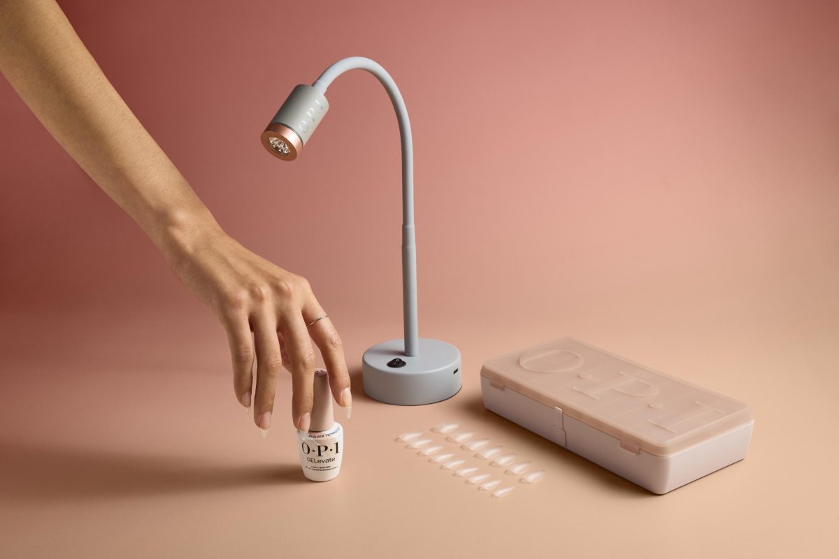 A hand holding a bottle of OPI GELevate nail polish is positioned next to a set of artificial nails, a nail lamp, and a nail kit box. The nail lamp has a flexible neck and a round base with a power button. The background is a gradient of pink and peach tones.