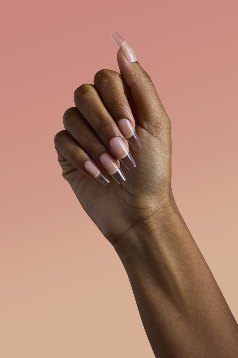 A close-up image of a hand with well-manicured, long, clear, and glossy nails. The nails have a smooth finish and are carefully shaped. The background is a gradient of soft pink and peach tones, complementing the aesthetic appeal of the manicure.