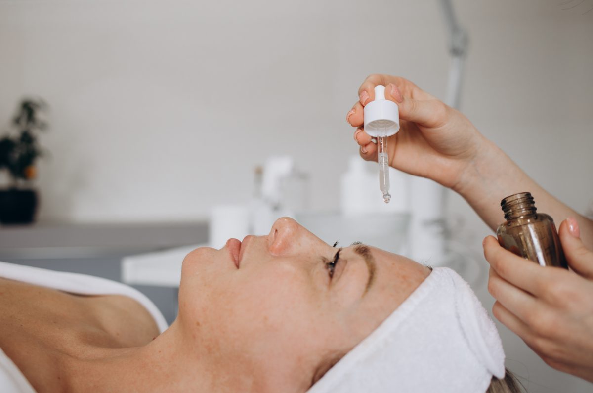 Cosmetologist applying few drops of moisturizing oil with a pipette on face her young pretty woman at spa center. Procedure for facial skin care