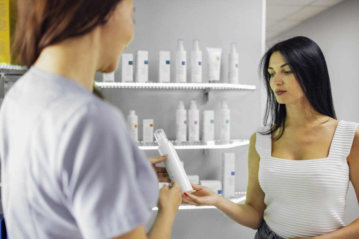 Saleswoman demonstrating bottle of cream with vitamins to female customer during work in beauty shop