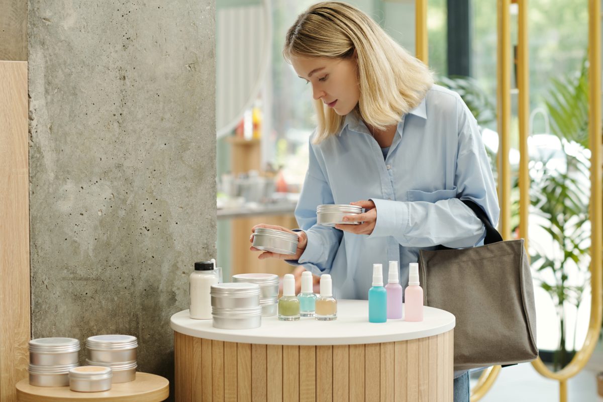 Young blond woman choosing body cream or scrub in tin jar on display with various beauty care products in cosmetic supermarket. Nail Care Retail Kits for Salons.