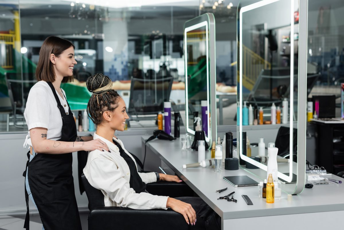 Top Hair Salon Brands Used by Professionals. client satisfaction, salon experience, happy hair stylist and female client with braids looking at mirror, cheerful women, hair bun, customer in salon, beauty service, tattooed, side view.