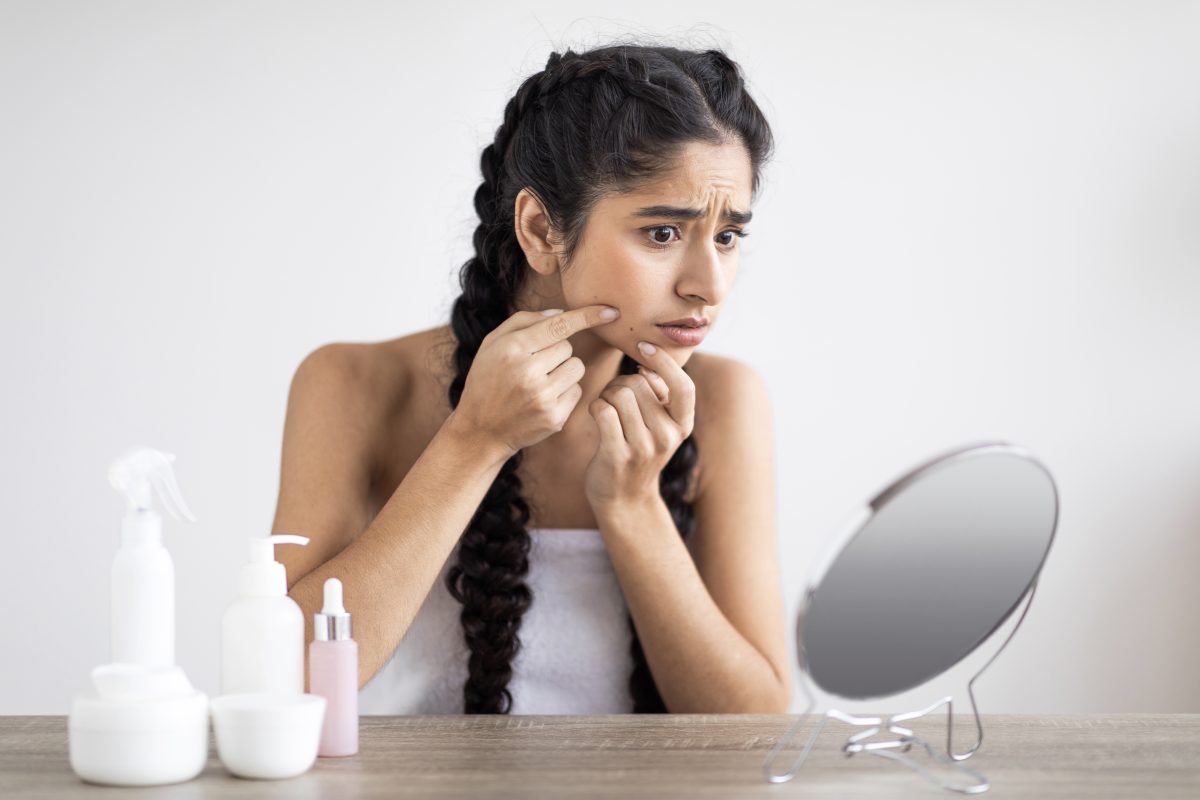 Upset Indian Woman With Problem Skin Looking At Pimple On Cheen While Sitting In Front Of Mirror At Home, Worried Young Eastern Female Suffering Acne While Making Daily Beauty Routine