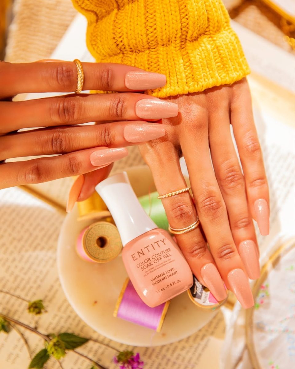 Freshly manicured hands with glossy peachy-nude coffin-shaped gel nails from Entity Spring 2025 collection. Styled with gold rings and a yellow knit sweater, the hands rest near a dish containing ENTITY Color Couture gel polish in 'Vulnerable Love,' surrounded by colorful thread spools, flowers, and an open book for a cozy, artisanal vibe.