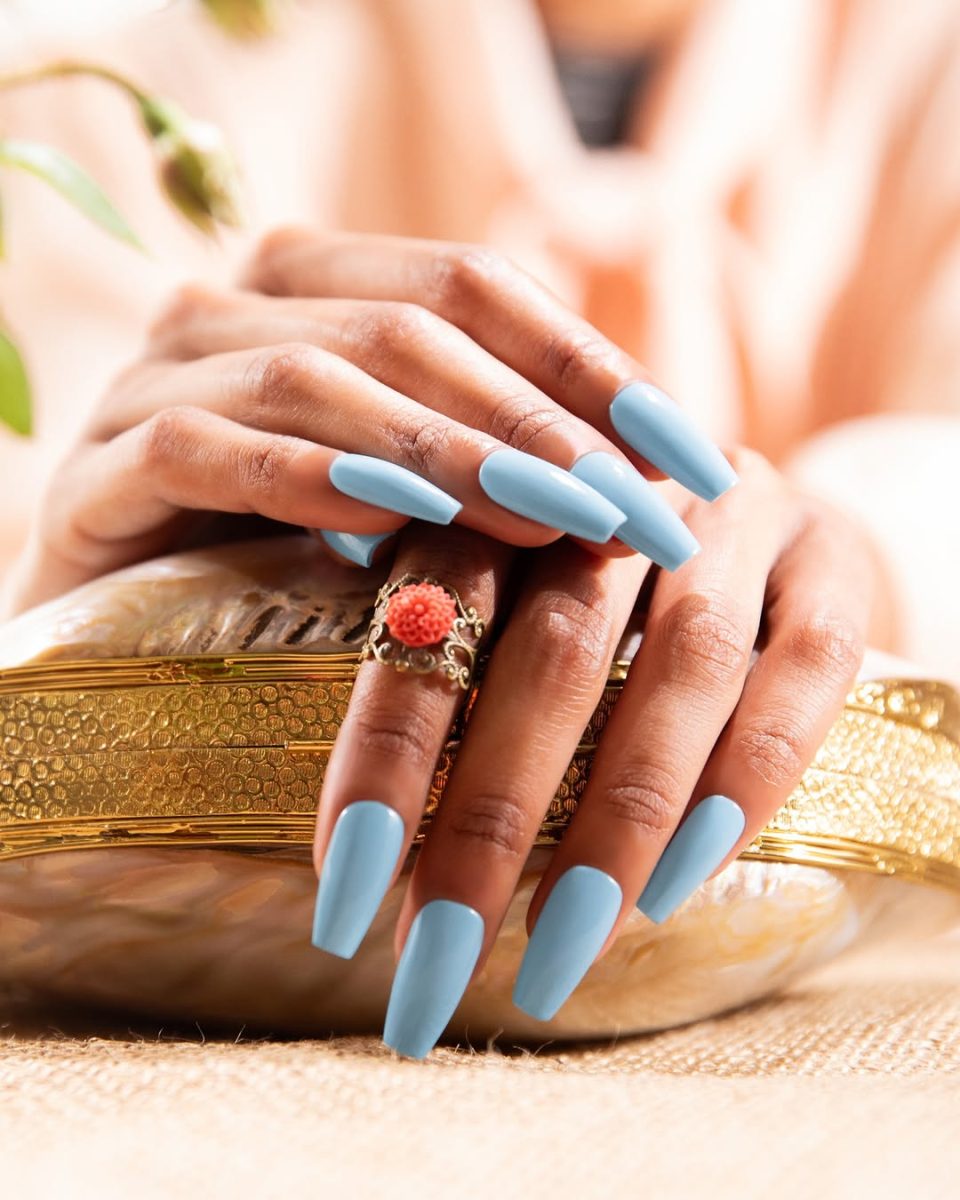 Close-up of hands with long, square-shaped matte light blue acrylic nails from Entity Spring 2025 collection, posed over a textured gold-accented clutch. One finger features a red floral statement ring, adding contrast and elegance. The soft-focus background enhances the luxurious and fashion-forward aesthetic.