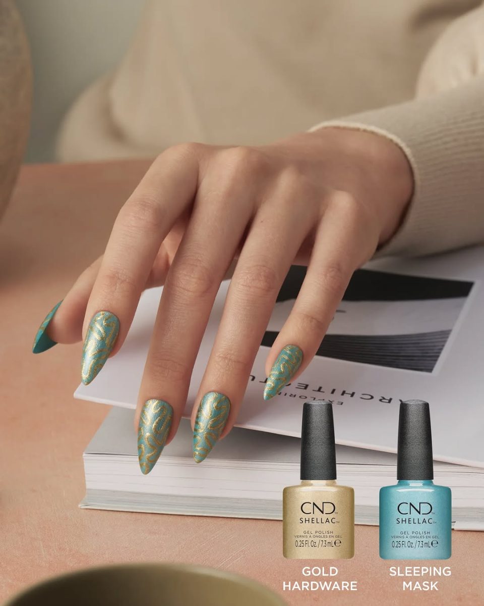 A close-up of a hand with long, intricate nail art using CND Shellac 'Sleeping Mask' (blue) and 'Gold Hardware' (gold shimmer), a key look from the Quiet Luxury Spring 2025 collection, with both polish bottles shown on a book.