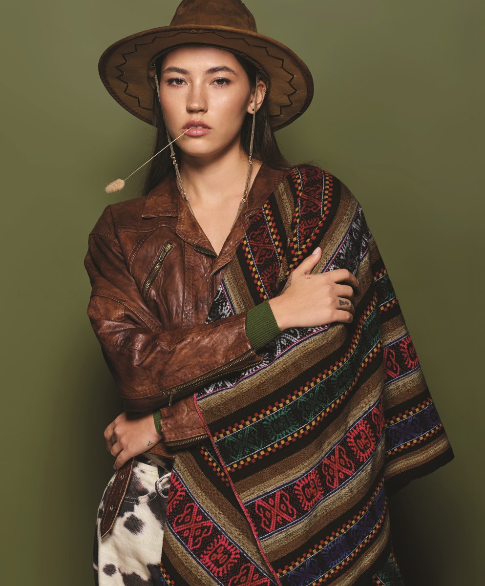 Model styled in Western-inspired fashion with a brown leather jacket, wide-brimmed hat, and vibrant geometric textile, evoking the earthy, artisanal spirit of CND Fall 2025 nail color collection. The olive-green backdrop and rustic styling reflect the season’s embrace of heritage tones, textured layers, and bold individuality.