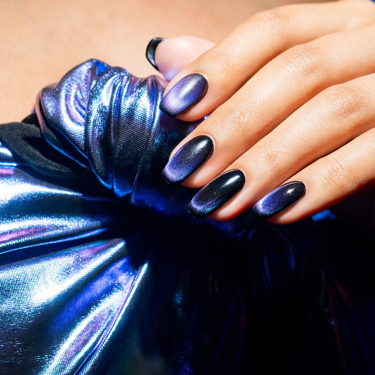 Close-up of manicured nails featuring Gelish Cat Eye polish in Blue Eclipse, with a glossy purple-black gradient and shimmering magnetic effect, resting on iridescent blue and purple fabric — perfect for nail art inspiration and salon marketing. Spring nails. Spring 2025 nails.