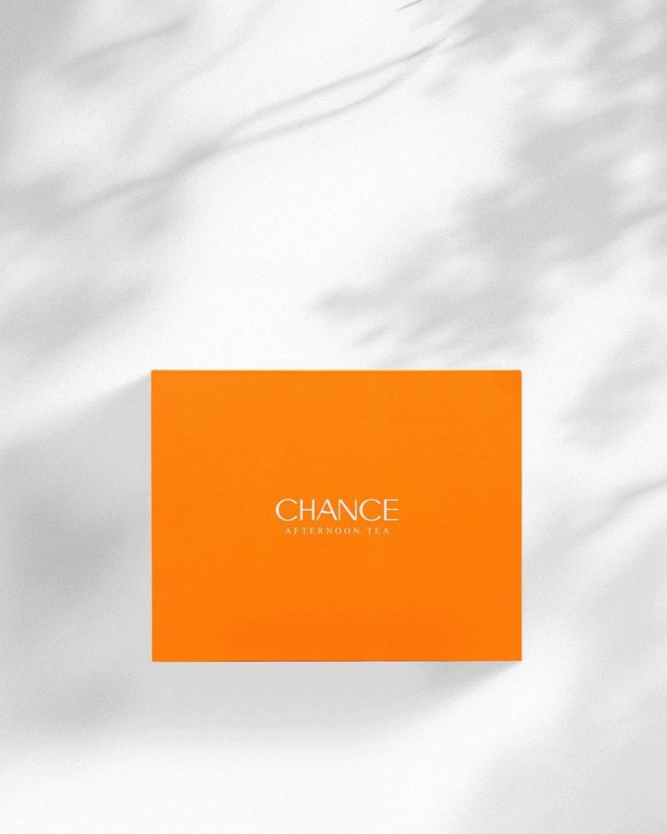 A minimalist, overhead shot of the closed Chance "Afternoon Tea" gel polish collection box. The box is a solid, vibrant orange with the words "CHANCE AFTERNOON TEA" printed in white centered on the lid. Dappled shadows fall across the image from the top.