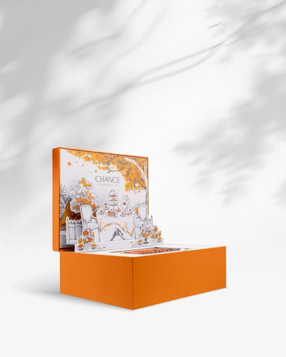 The Chance "Afternoon Tea" Gel Collection display box is shown at an angle, resting on a white surface with dappled shadows. The signature orange box base is beneath the lid, which stands up to reveal the black-and-white illustration of the elegant autumn garden tea party scene framed by orange fall leaves. Chance Creation Collections.